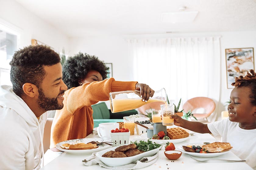 family in a casual bright dining area, gathered around a breakfast table with sausage patties, waffles, berries, and juice