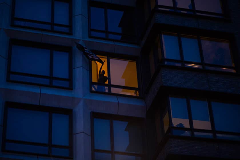 a building in the twilight, with the orange glow of an interior light around a figure holding a flag in a window