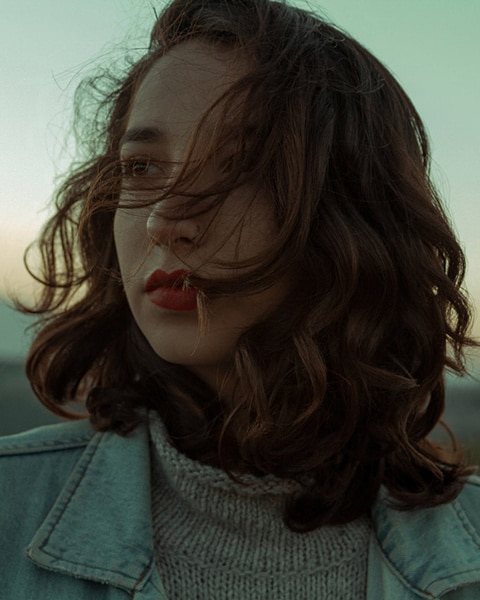 headshot of a young woman wearing red lipstick and looking to the left of the frame, her hair blowing across her face