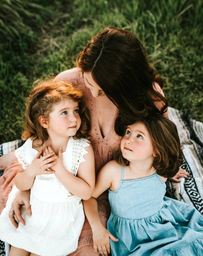 mother with two daughters sitting partially on her lap and leaning into her chest, looking up at her face